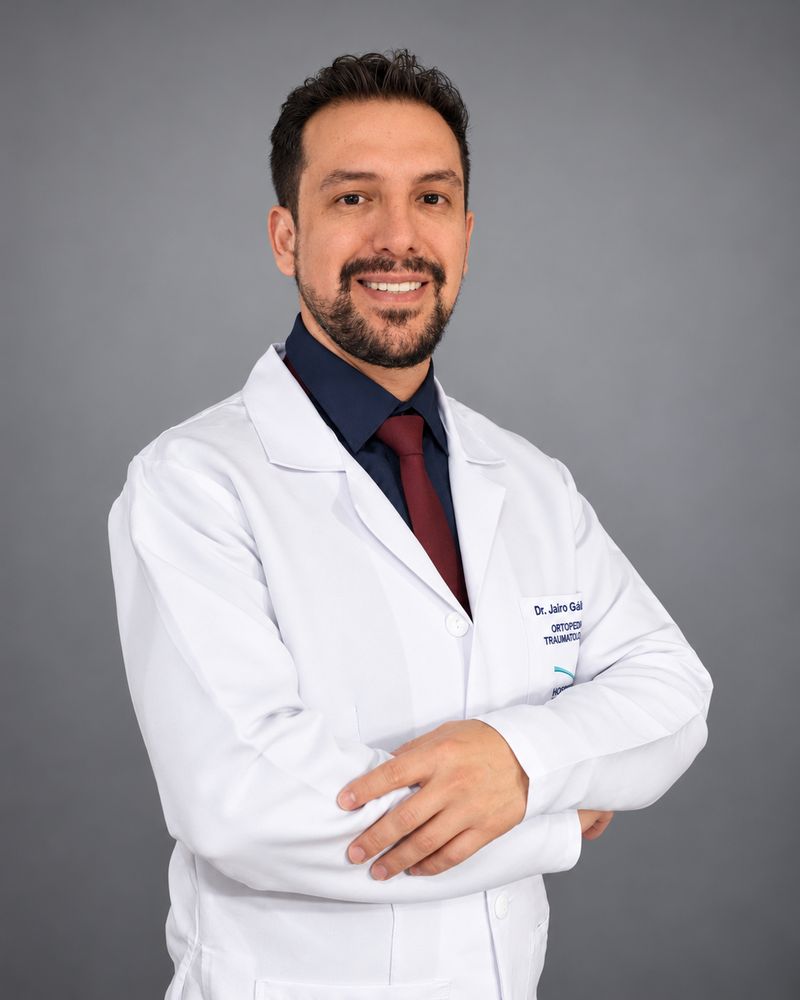 El Dr. Jairo Gallardo, traumatólogo especialista en rodilla, sonriendo con bata médica y los brazos cruzados.